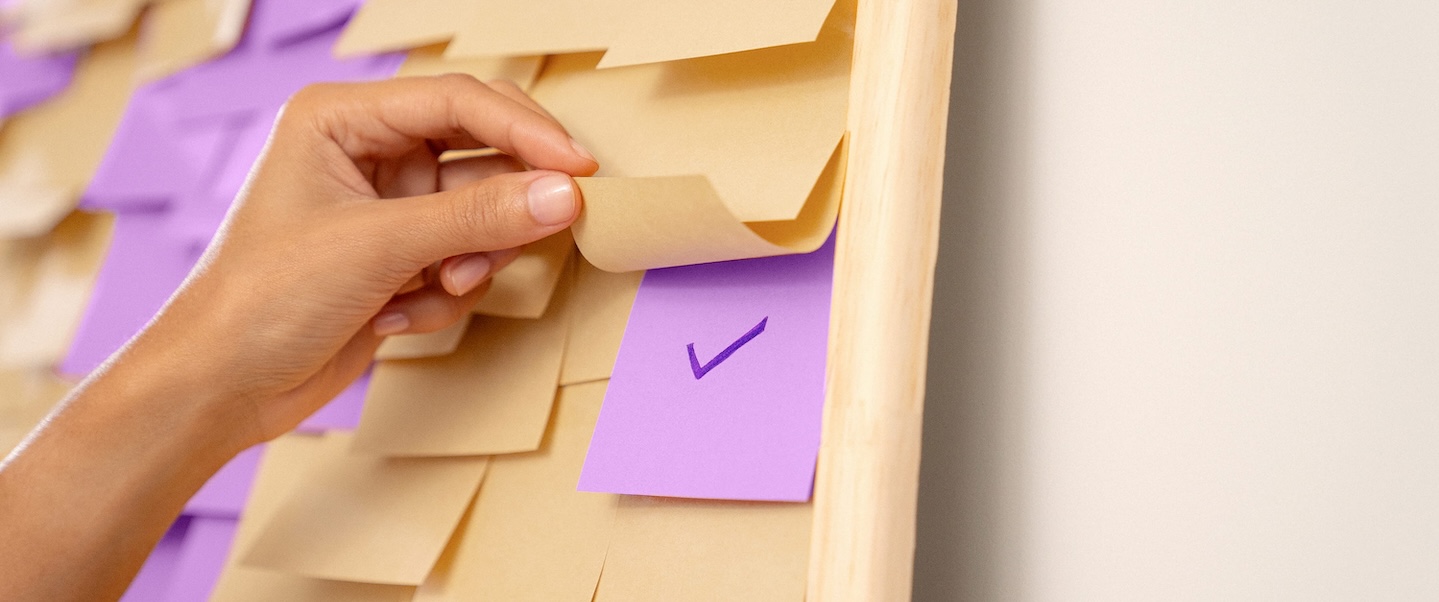 Uma mão branca segura um post-it amarelo que está em uma lousa cheia de post-its amarelos e roxos.