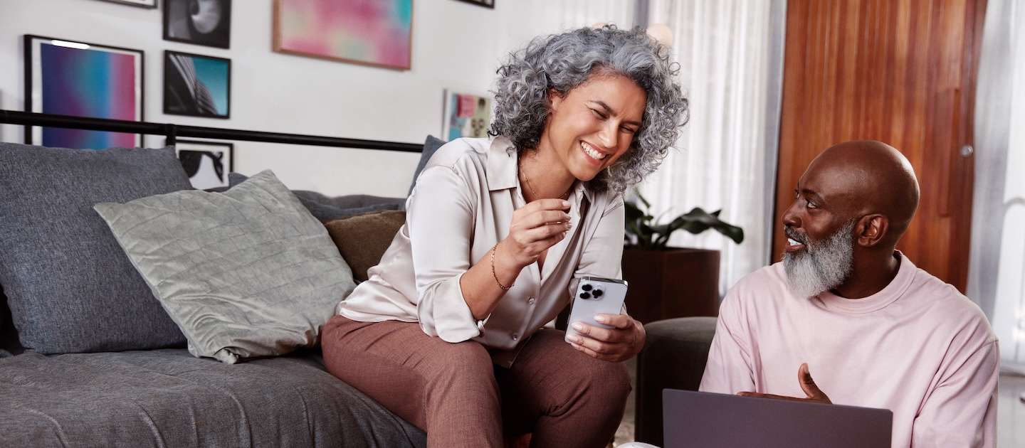 Mulher negra de cabelos cacheados e grisalhos sentada em um sofá e sorrindo, segurando com a mão esquerda um celular em direção a um homem negro, careca, com barba grisalha, sentado ao seu lado no chão, olhando para o rosto dela enquanto sorri e com uma pequena mesa de madeira a sua frente com um notebook.